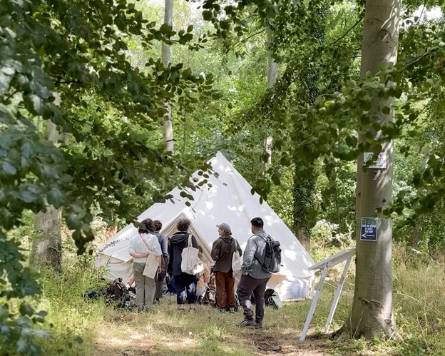 Participants gather near the entrance to a small white bell tent under the canopy of large green trees in a field of tall grass and flowers.